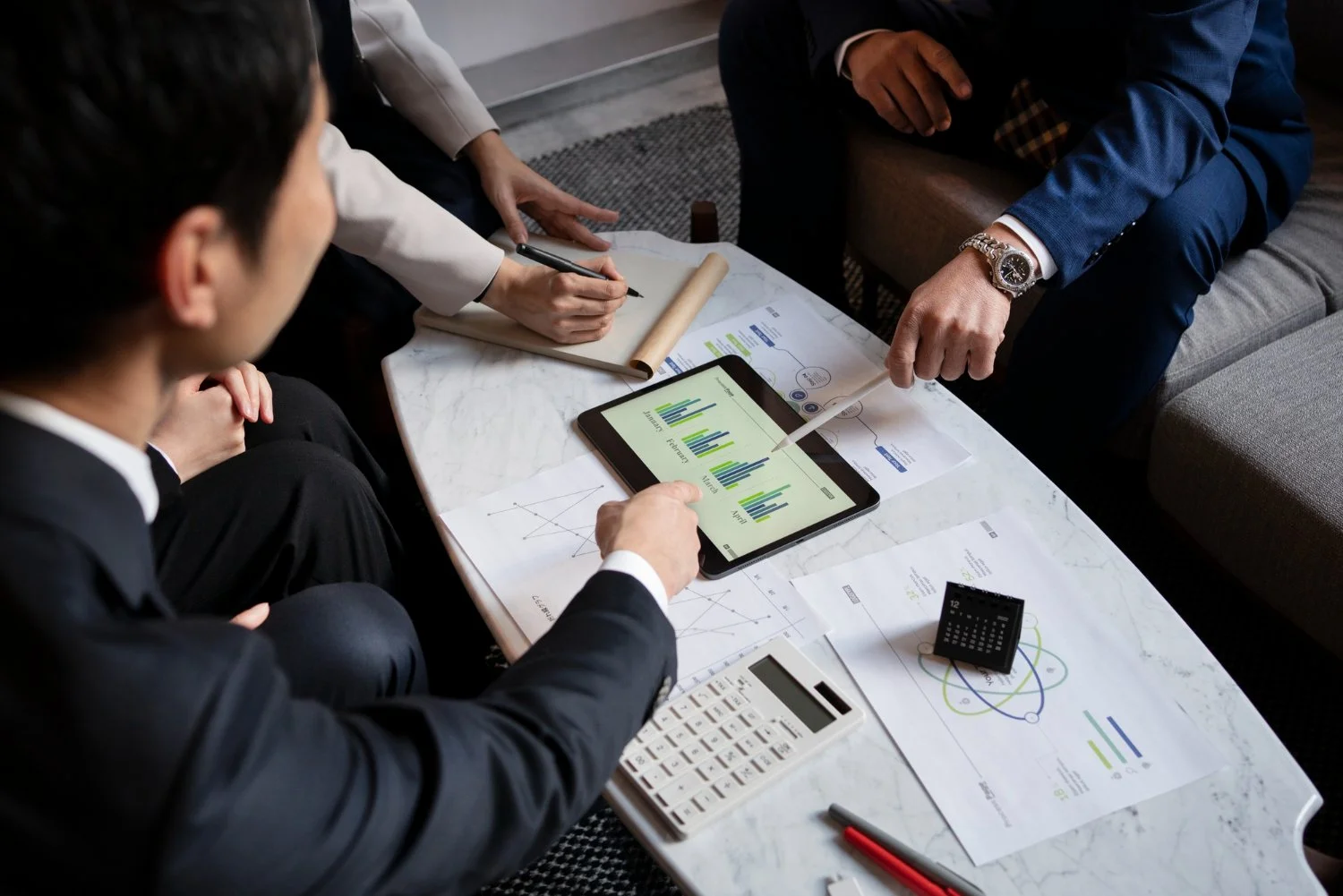 Business meeting with three people discussing charts and data on a tablet, with papers, calculator, and pen on a white table.